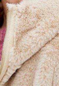 Beige fake fur jakke med en tekstureret, blød overflade. Har en cremet lynlås, synlige sømme og en bred krave.