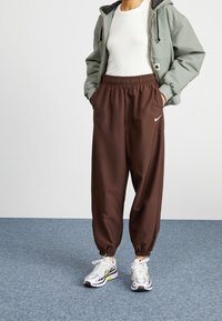 Hnědé Nike tepláky s elastickým pasem a manžetami, spárované se světle zelenou zipovou mikinou a bílými teniskami s šedými akcenty.