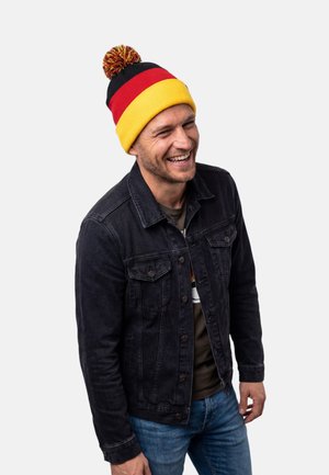 Mies hymyilee, hänellä on musta farkkutakki, siniset farkut ja pom-pom-pipo, jossa on mustia, punaisia ja keltaisia raitoja.