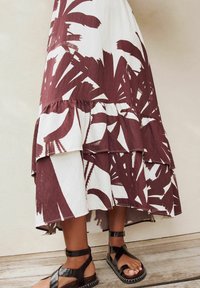 Maxikleid mit floralen Mustern in Burgunderrot und Weiß, ausgestattet mit Rüschenschichten. Getragen mit schwarzen Sandalen, die die Knöchelriemen betonen.
