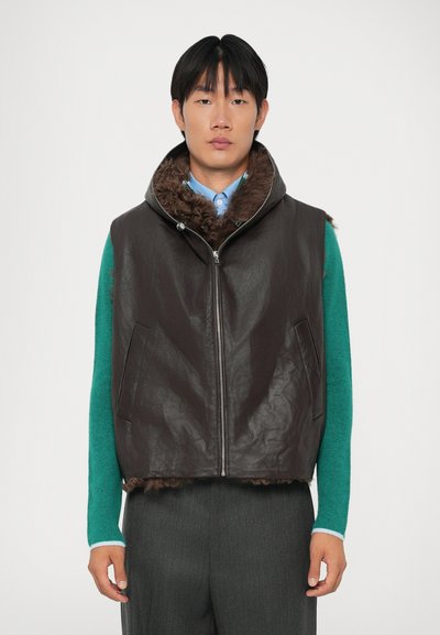 Uomo che indossa un gilet in shearling marrone scuro con zip sopra una maglietta a maniche lunghe teal e pantaloni grigi, in piedi su uno sfondo bianco semplice.