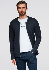 Dunkelblauer Strickcardigan mit Reißverschluss, gerippten Bündchen und glatter Textur, getragen über einem hellblauen bedruckten T-Shirt und dunklen Jeans.