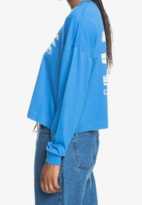 Niebieska krótka bluza z długimi rękawami, z nadrukiem logo na przodzie, o luźnym kroju i z miękkiego materiału. Noszona z dżinsami o wysokim stanie.