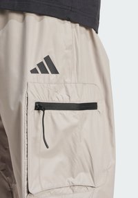 Beiga cargo-shorts i lättviktsmaterial, med en dragkedjeficka, synliga sömmar och en svart Adidas-logotyp på sidan.