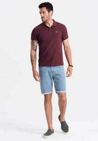 Viininpunainen polo-paita, jossa on kaulus ja rintalogointi, yhdistettynä vaaleansinisiin farkkushortseihin. Mallilla on päällään tummat liukupohja kengät, jotka korostavat rentoa tyyliä.