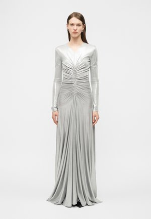 Nő hosszú ezüstfényű, fémes ruhában, elülső részén ráncolt részletekkel, hosszú ujjakkal és légiesen hullámzó szoknyával, egyszerű háttér előtt áll.