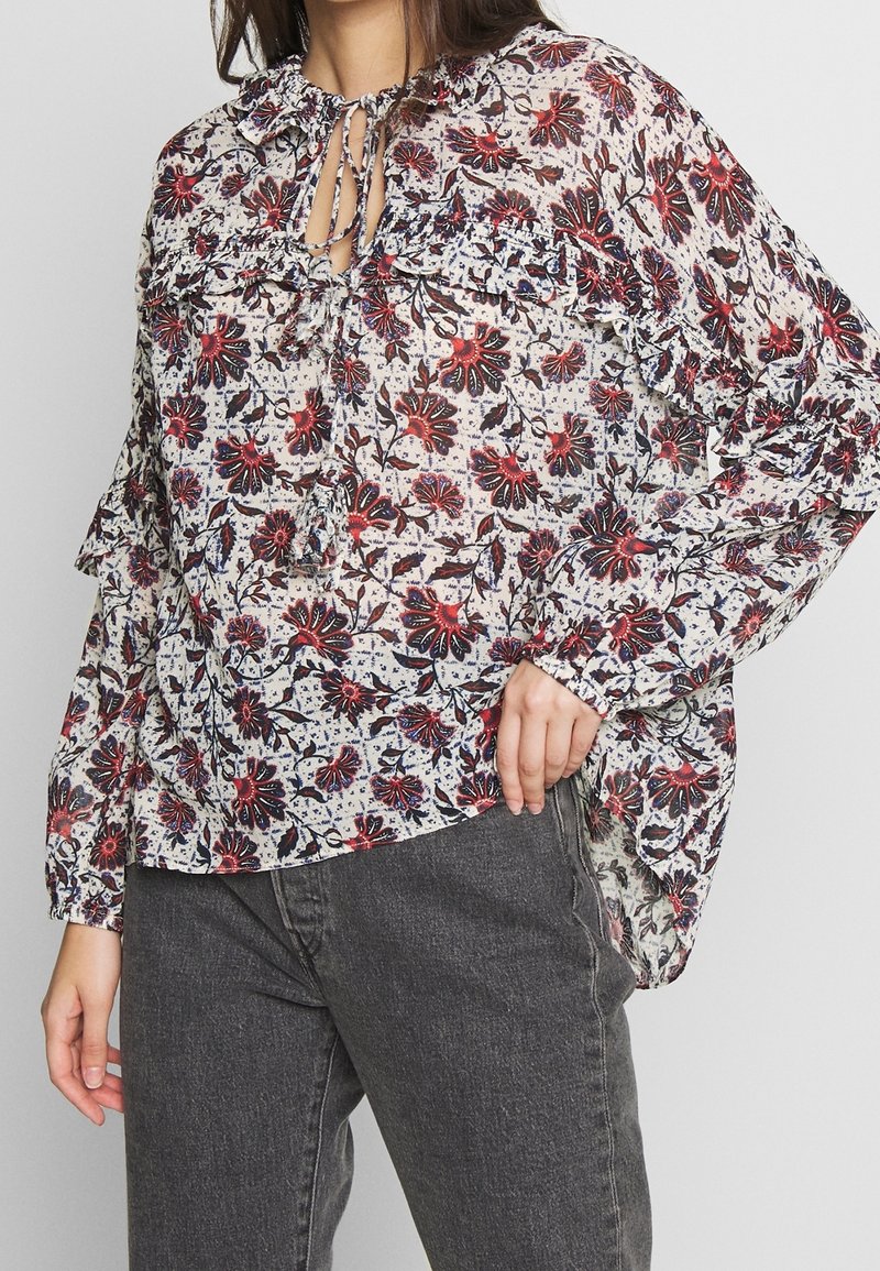 Blusa floreale in tessuto bianco trasparente con motivi di fiori rossi e blu, dettagli arricciati e chiusura con fiocco al collo. Abbinate a jeans grigi scuri.