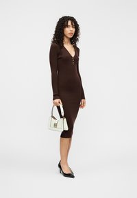 Καφέ φόρεμα με ριμπ ύφανση, V-λαιμόκοψη και λεπτομέρειες με κουμπιά, συνδυασμένο με λευκή δομημένη τσάντα χειρός και μαύρες μυτερές γόβες.