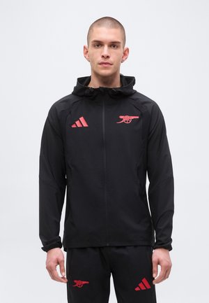 Mężczyzna ubrany w czarną sportową kurtkę i spodnie z czerwonymi logo Arsenal i Adidas, stojący na jednolitym jasnym tle.