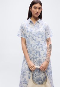 Φόρεμα κοντών μανικιών σε ανοιχτό γαλάζιο με κουμπιά μπροστά, διακοσμημένο με λεπτομερές μπλε σχέδιο δέντρων και μορφών. Το μοντέλο κρατάει μια μπεζ τσάντα.