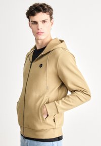 Béžová mikina na zips s čiernym logom, šnúrkami, bočnými vreckami a rebrovaným lemom. Nosená cez čierne tričko a modré džínsy.