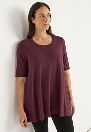 Woman wearing a loose-fitting, short-sleeve, maroon top and black pants, standing against a white wall and looking to the side.
