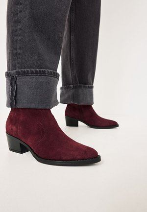 Person iført mørke op rullede jeans og bordeaux ruskind ankelstøvler med lave blokhæle på et hvidt gulv.