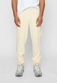 Lys beige cargo bukser med elastisk midjebånd, sidelommer og avsmalnede mansjetter, kombinert med grå joggesko. Glatt stofftekstur.