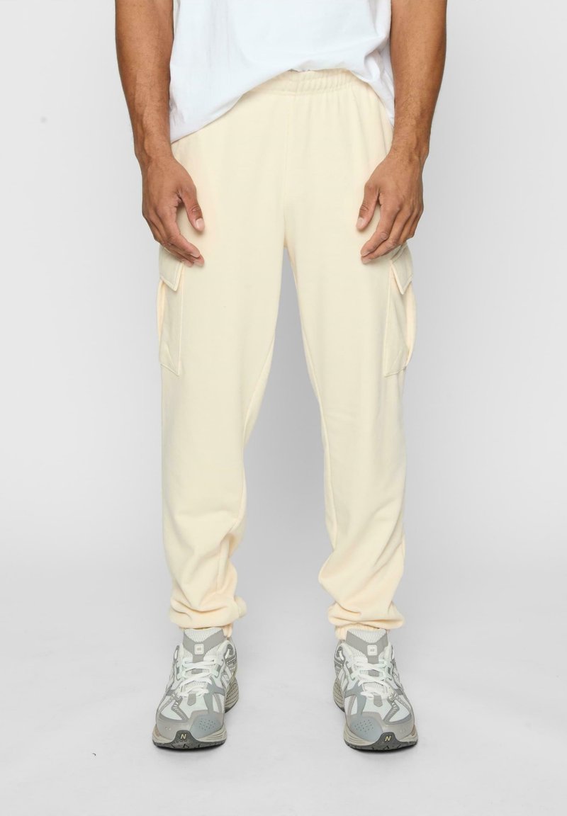 Pantalons cargo beige clair avec taille élastique, poches latérales et chevilles fuselées, associés à des chaussures de sport grises. Texture de tissu lisse.