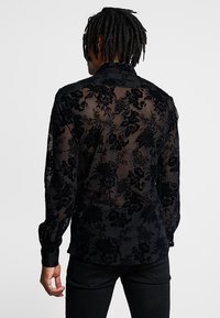 Camicia nera in pizzo floreale con design trasparente, caratterizzata da maniche lunghe e collo a camicia. Il tessuto presenta un aspetto testurizzato e a motivi.