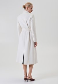 Donna con un chignon biondo che indossa un lungo cappotto bianco con cintura e tacchi a slitta scuri, in piedi e che guarda oltre la spalla contro uno sfondo semplice.