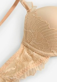 Bežinis liemenėlė su nėrinių detalėmis, minkštais paminkštintais puodeliais ir reguliuojamomis petnešomis, su sudėtingais gėlių raštais nėriniuose ir lygiu audiniu.