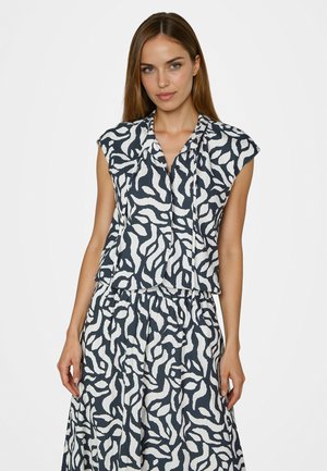 Frau trägt ein ärmelloses Top mit schwarz-weiß abstraktem Muster und passenden Rock vor einem einfarbigen weißen Hintergrund.