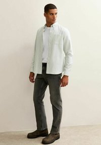 Camicia a righe verde chiaro e bianca con taschino sul petto, abbinata a jeans grigi e calzature nere. Tessuto di seta morbida.