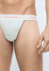 Svetlozelený thong Calvin Klein s úzkym pásom, na ktorom sa nachádza koralová značka. Hladká textúra, priliehavý strih zdôrazňujúci tvar tela.