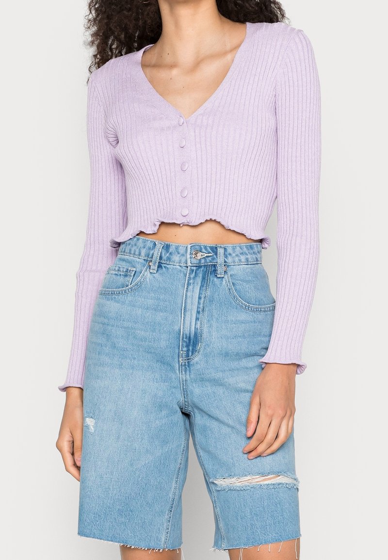 Světle fialová žebrovaná zkrácená kardigán s výstřihem do V a zapínáním na knoflíky, kombinovaná s modrými džínovými šortkami s poškozenými detaily.