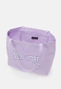 Sac fourre-tout en toile lilas avec le texte "NEW YORK" en blanc et or, doté de deux poignées et d'un design simple. L'intérieur n'est pas doublé et comporte une étiquette.