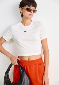Balta trumpa t-marškiniai su juodu Nike logotipu, derinama su oranžinėmis sportinėmis kelnėmis. Modelis laiko juodą rankinę ir dėvi žalius katės akies stiliaus akinius.