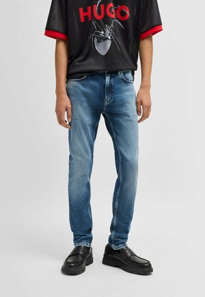 Jeans in denim blu con una vestibilità slim, caratterizzati da un lavaggio sbiadito e un classico design a cinque tasche. Abbinati a una t-shirt nera con grafica e scarpe nere.