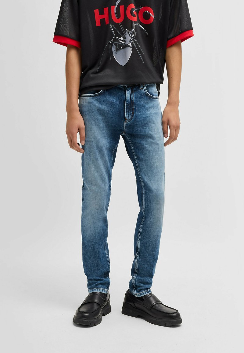 Jeans in denim blu con una vestibilità slim, caratterizzati da un lavaggio sbiadito e un classico design a cinque tasche. Abbinati a una t-shirt nera con grafica e scarpe nere.
