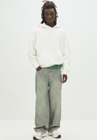 Weiße Kapuzenjacke, grün gestreiftes Hemd darunter, hellblaue weite Jeans und silberne Sneaker. Das Model steht vor einem schlichten Hintergrund.