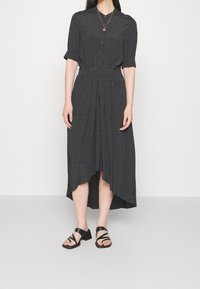 Kobieta w czarnej midi sukience w białe kropki, z półrękawami, z guzikami z przodu oraz czarnych sandałach na niskim obcasie z paskami.