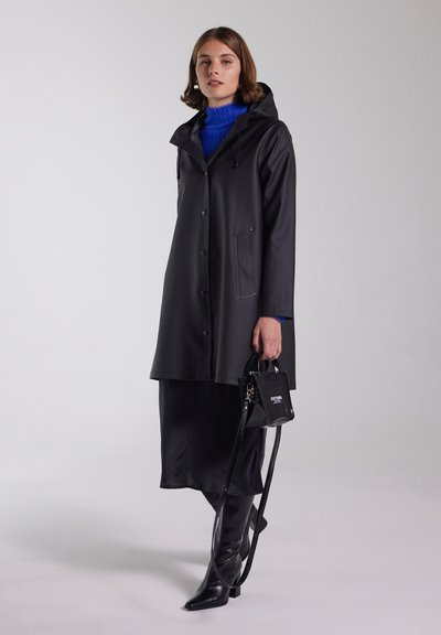 Svart vattentät kappa med huva, tryckknappar, två framfickor, bärs över en blå tröja och svart kjol, kombinerad med en liten svart handväska.