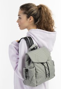 Zaino grigio trapuntato con flap pieghevole, spallacci neri e dettagli con cordoncino. Presenta cuciture diagonali e una superficie testurizzata.