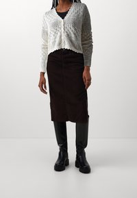 Vit stickad kofta med ett diamantmönster, knäppning framtill; ihop med en brun kordflöjels midi-kjol. Svarta knähöga stövlar fulländar looken.