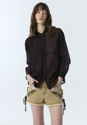 Joven mujer que lleva una camiseta negra transparente de manga larga y pantalones cortos de mezclilla beige con bordado floral, de pie con las manos en los bolsillos.