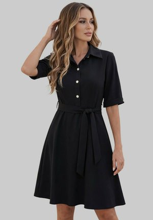 Femme aux cheveux longs et ondulés portant une robe noire boutonnée avec des manches aux coudes et une ceinture nouée à la taille, regardant sur le côté.