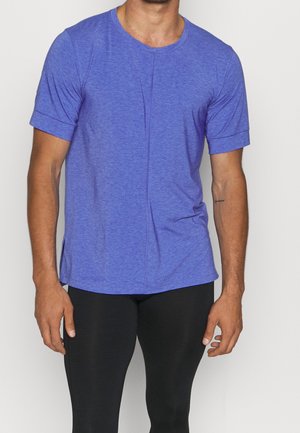 Man draagt een nauwsluitend, paars gemêleerd T-shirt met korte mouwen en zwarte legging, staand tegen een eenvoudige lichte achtergrond.