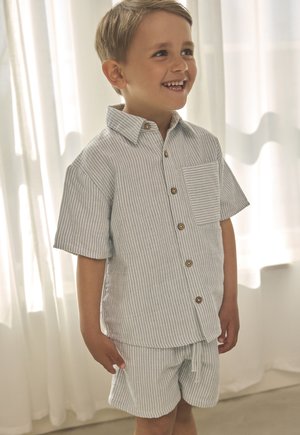 Lachende jongen staat binnen, gekleed in een lichtblauw-wit gestreept shirt met korte mouwen en bijpassende korte broek met knopen en zakdetails.