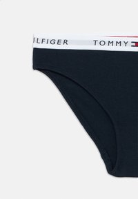 Tumesinine puuvillane bikiinipüksik, millel on elastne vöökoht valge triibu ja punaste detailidega. Logotekst vöökohal. Sujuv tekstuur.