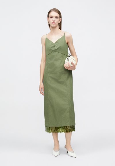 Γυναίκα με μίντι φόρεμα σε χακί πράσινο χρώμα χωρίς μανίκια και φράντζα στο τελείωμα, κρατώντας έναν λευκό φάκελο και φορώντας λευκές μυτερές γόβες, σε απλό φόντο.