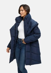 Marineblauwe gewatteerde jas met een hoge kraag, doorgestikte ontwerp, ritsluiting en zijknopen. Gecombined met een witte top en donkere jeans.