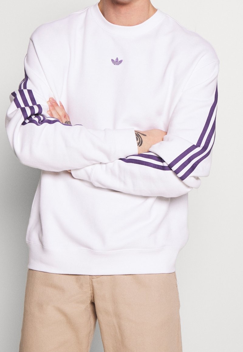 Vit sweatshirt i bomullsmix, med lila adidas-logotyp och tre lila ränder ned längs varje ärm. Crew neck och ribbade muddar.