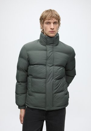 Joven con cabello claro que lleva una chaqueta de invierno acolchada de cuello alto de color verde oscuro y pantalones negros, de pie frente a un fondo sencillo.
