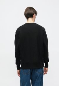 Μαύρο φούτερ με �στρογγυλή λαιμόκοψη, ριμπ μανσέτες και τελείωμα. Το ύφασμα φαίνεται μαλακό με ελαφρώς υφή στην επιφάνεια. Φορώντας με μπλε τζιν παντελόνι.