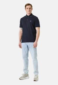 Polo shirt bleu marine en tissu texturé et à manches courtes, doté d'un col et d'un petit emblème. Accompagné d'un jean bleu clair et de baskets grises.