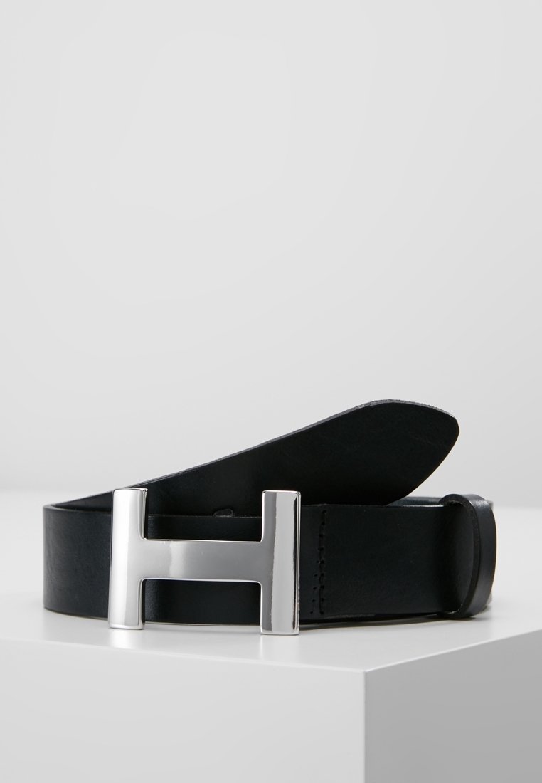 Ceinture en cuir noir avec une grande boucle métallique argentée en forme de lettre H, posée sur une surface blanche.