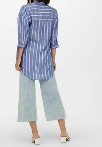 Camicia lunga a righe blu e bianche con maniche arrotolate, abbinata a pantaloni in denim a gamba larga di un leggero blu e sandali bianchi con tacco a blocco.