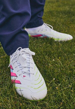 Zapatillas deportivas blancas con superficies texturizadas, acentos en verde y rosa, y un patrón distintivo en la suela, colocadas sobre hierba.