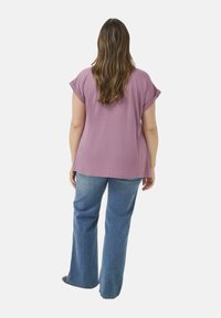 Žena s dlouhými hnědými vlasy, oblečená v volném mauve krátkém topu s krátkým rukávem a modrých rozšířených džínách, stojící zády k objektivu na bílém pozadí.